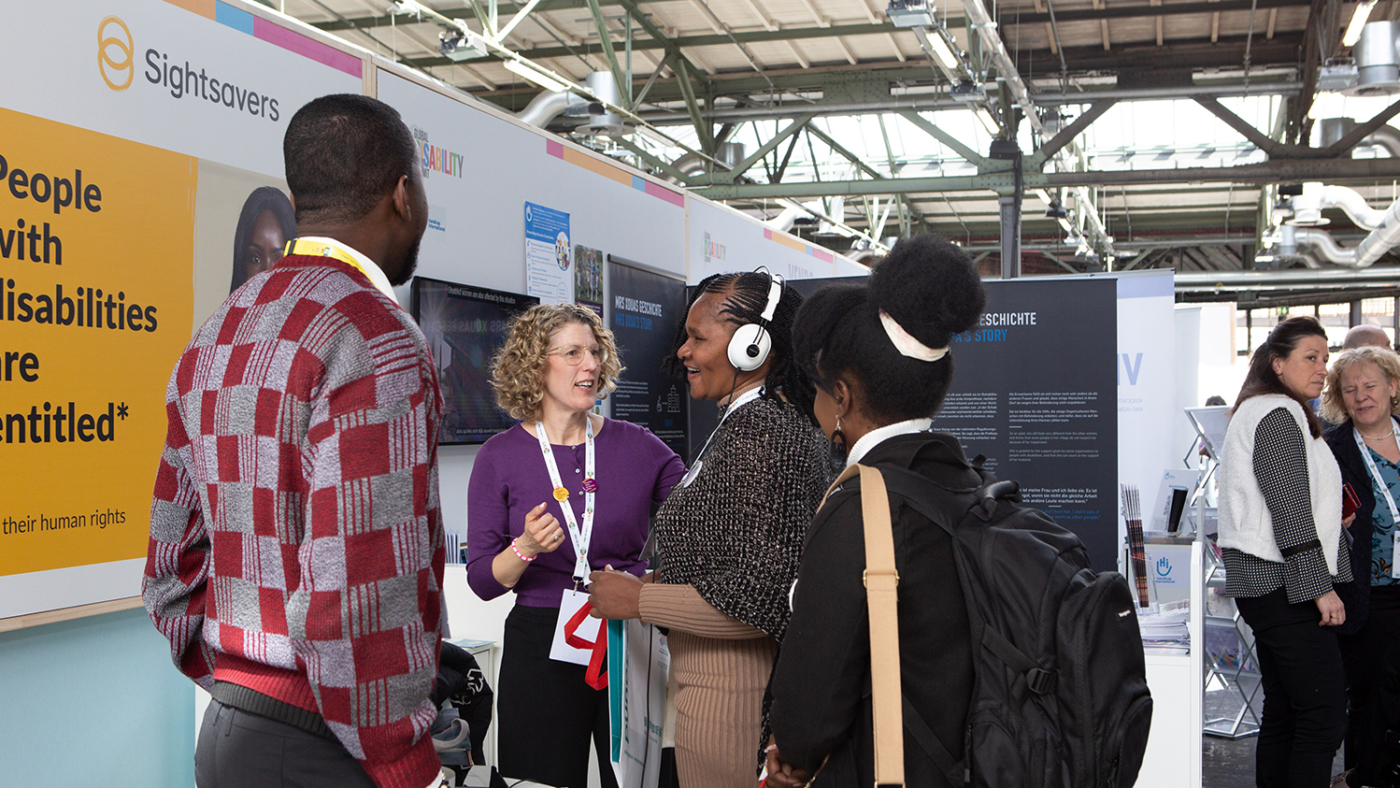 A group of people gather at the Sightsavers' booth. One person is wearing headphones.
