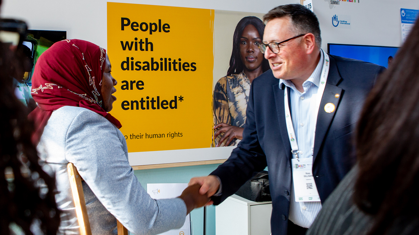 Ross, who wears a suit, shakes hands with Ummy at the Sightsavers-branded booth.