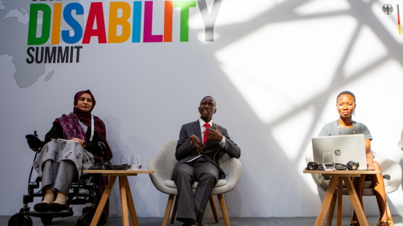 Three panellists are seated with the Global Disability Summit logo behind them. One woman in a wheelchair sits beside a young man wearing a suit.