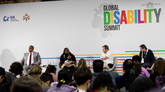 An audience watches a panel with the Global Disability Summit backdrop behind them.