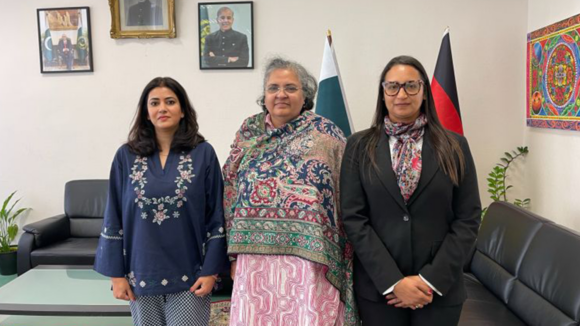 Three women stand next to each other smiling, with flags and paintings in the background.