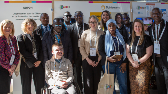 A large group of people smile in front of a Sightsavers-branded booth.