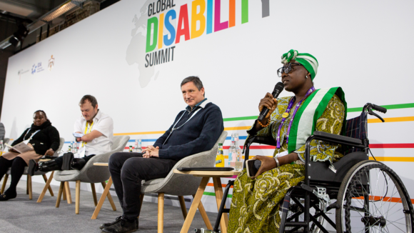 A woman in a wheelchair speaks into a microphone alongside a panel. The Global Disability logo is in the background.
