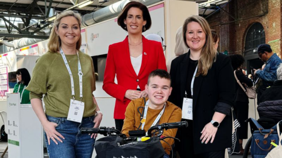 Three smiling women stand behind a young boy on a mobility scooter.