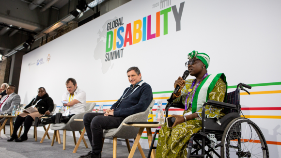 A woman in a wheelchair speaks into a microphone alongside a panel. The Global Disability logo is in the background.