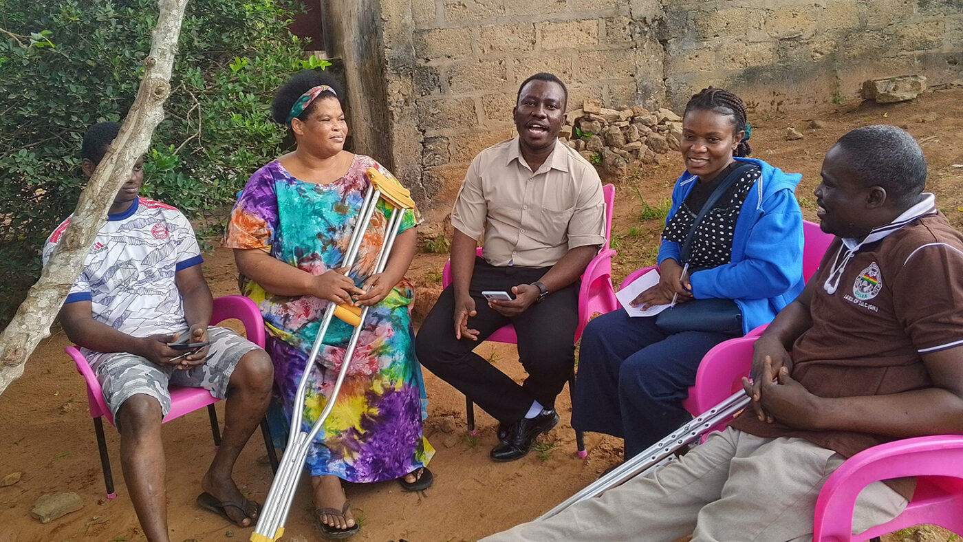 A group of five people in conversation sit in a semicircle under a tree during a qualitative interview.