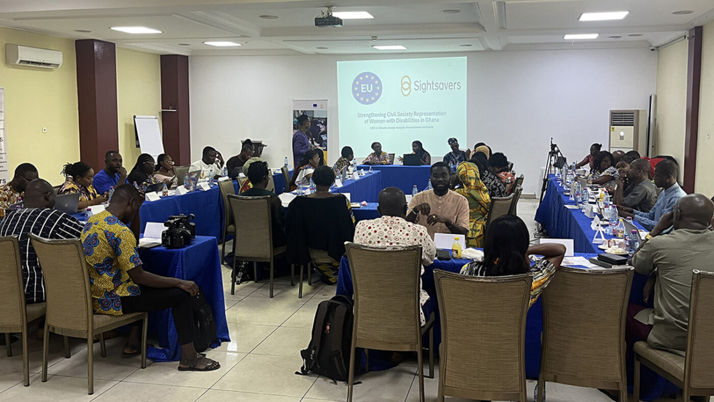 In a large meeting room, people sit around tables watching a presentation on the far wall.