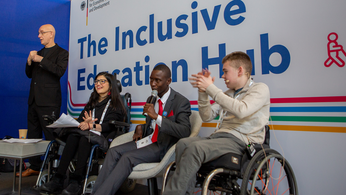 A man with a microphone sits in between two other panellists in wheelchairs. Large lettering saying 'The Inclusive Education Hub' can be seen behind them.