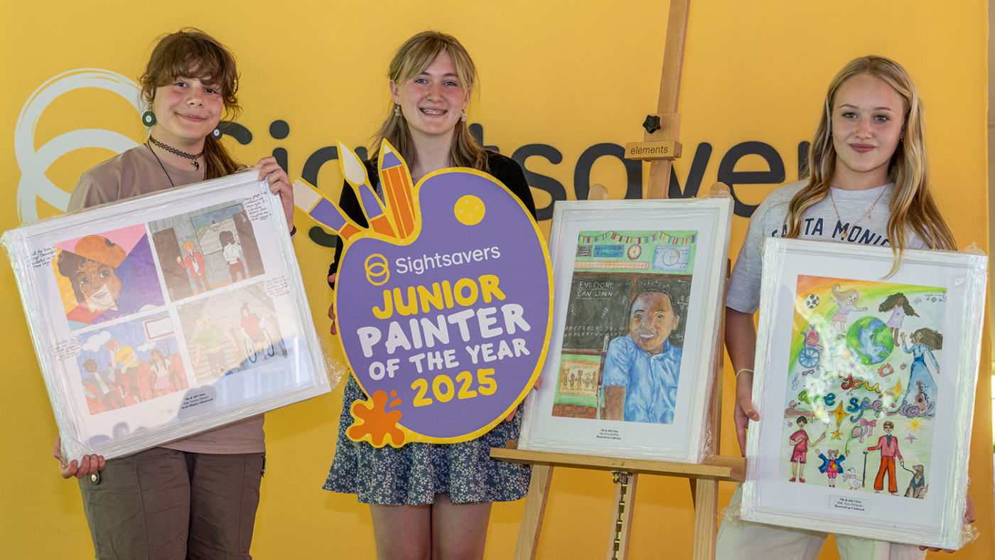 Two girls hold their artwork while another holds a 'Junior Painter of the Year' sign next to an easel and canvas.