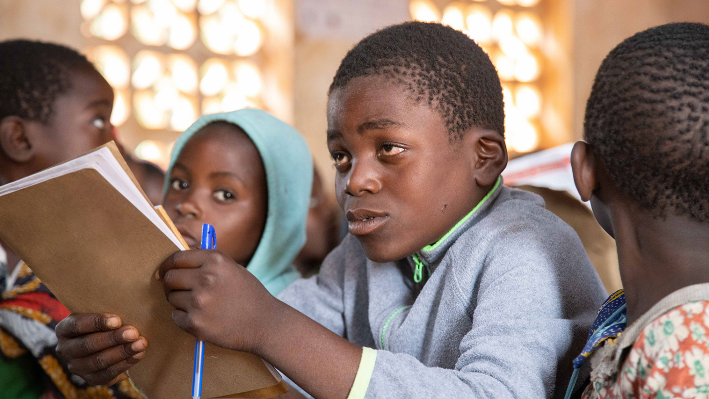 Chisomo is concentrating as he sits at a classroom desk holding a pen and notepad.