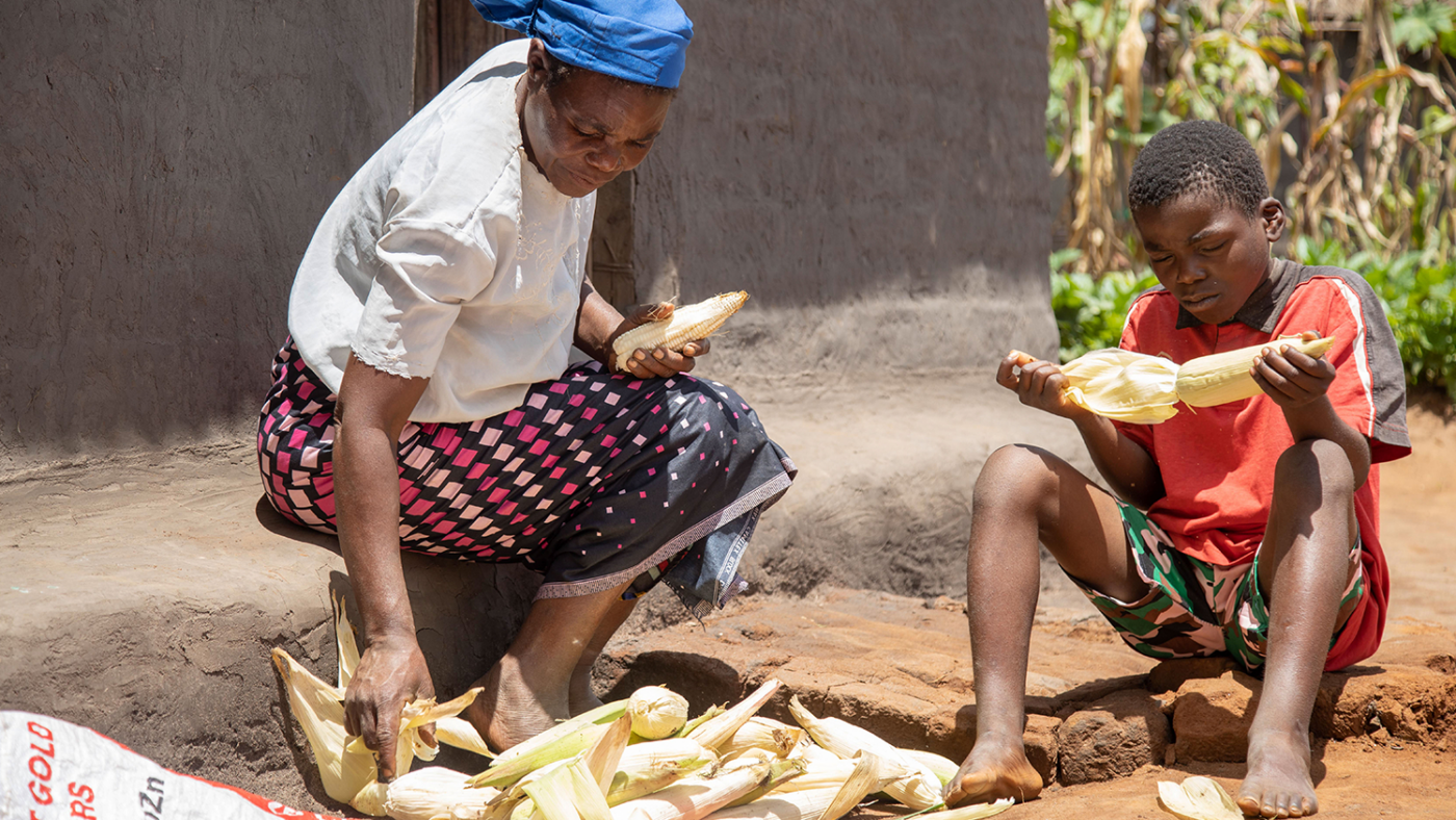 Chisomo and his mother sit on the ground as they prepare corn cobs.