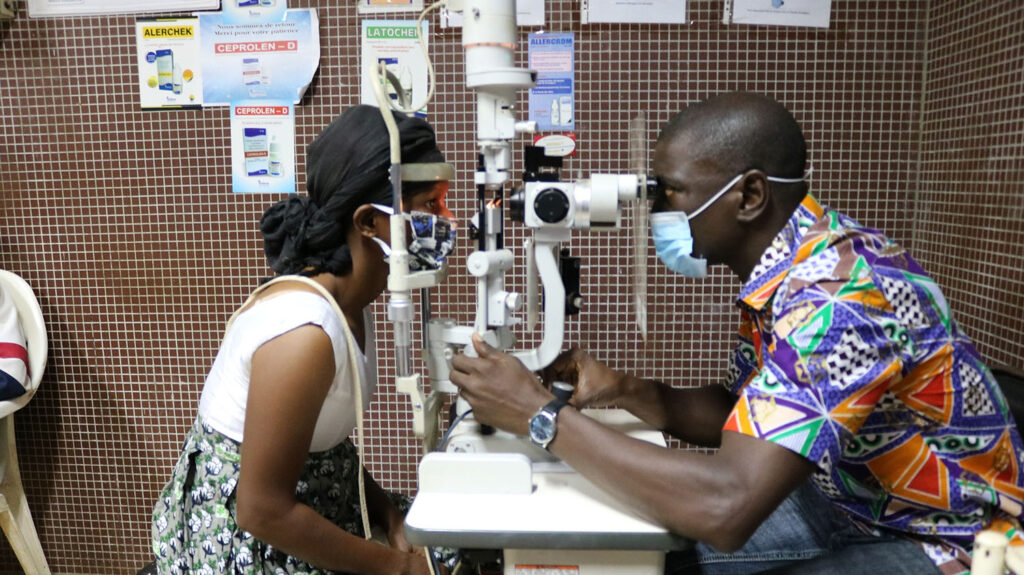 An eye health worker examines a woman's eyes for signs of trachoma. They're both wearing medical masks.