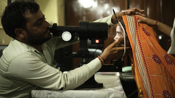 A man holds a woman's face in position while using a medical instrument to examine her eyes.