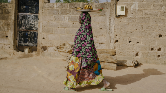 A woman in traditional African dresswear walks along a street while balancing a tray of fruit on her head.