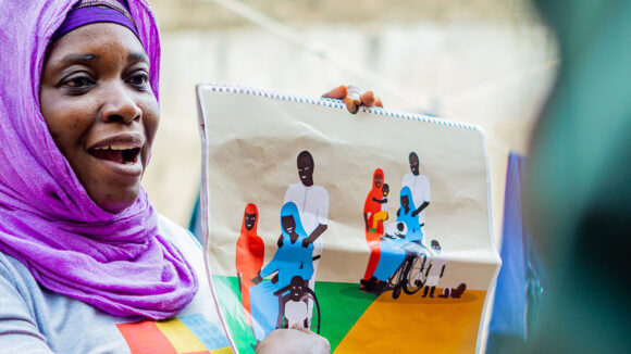 A woman wearing a hijab holds up an illustration of a family, in which the mother is using a wheelchair.