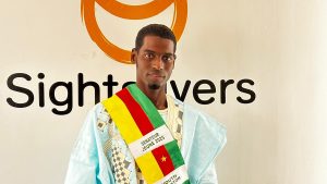 Ousmanou is wearing his youth senator sash in the colours of the Cameroon flag. Behind him on a wall is the Sightsavers logo.