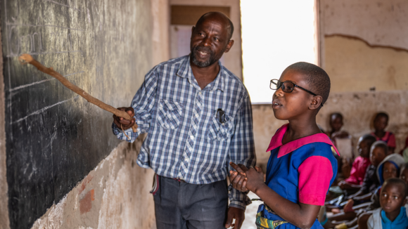 Melia is wearing glasses while standing at the blackboard with her male teacher. Her teacher is pointing to a word on the blackboard.