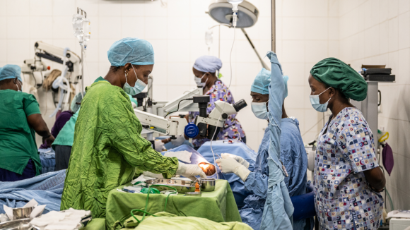 Five medical professionals stand around an operating theatre during a cataract operation. They are all wearing medical masks and medical scrubs.