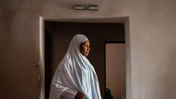 A woman wearing a hijab stands in the doorway at a hospital. Above the door, the sign reads 'Family planning unit'.