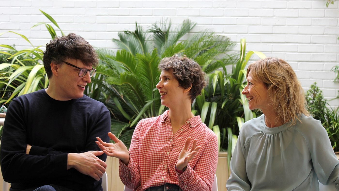 A man and two women talk while sitting in an outdoor space. There are lush, green plants behind them.