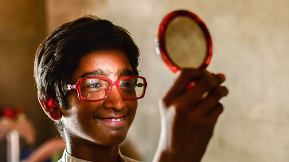 A boy holds up a mirror to check his reflection. He is smiling broadly and wearing a pair of glasses with a red frame.