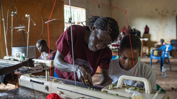 A woman leans over to fix a knitting loom while another woman, who is seated, watches.