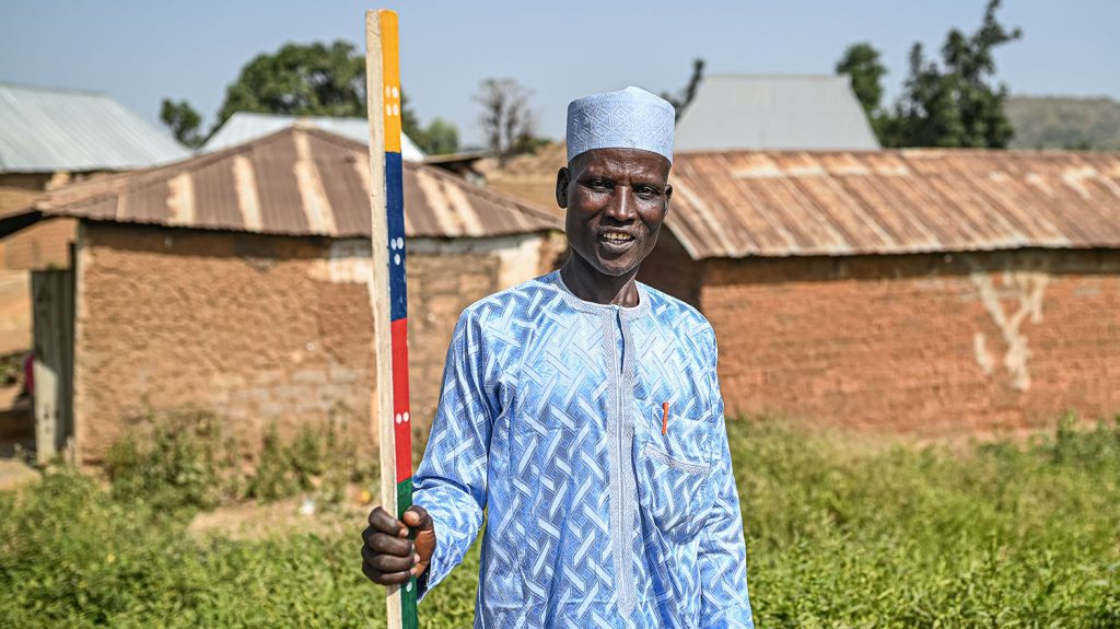 A male community drug distributor stands in a field holding a dose pole. He is wearing a blue tunic and cap.