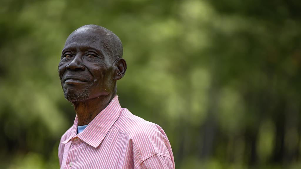 A man wearing a smart shirt smiles with greenery behind him.