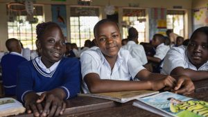 Three students sit a desk in a classroom wearing school uniform. One of the children has a physical disability.