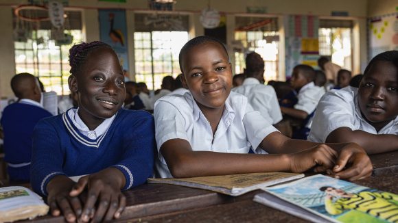 Three students sit a desk in a classroom wearing school uniform. One of the children has a physical disability.