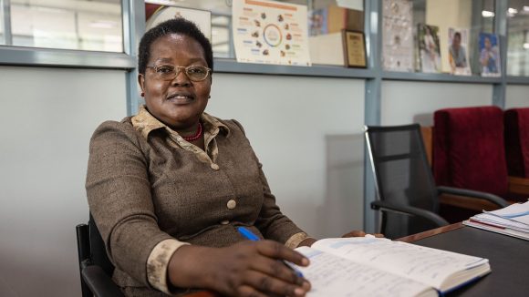 A woman wearing office clothes sits at a desk writing in an open book.
