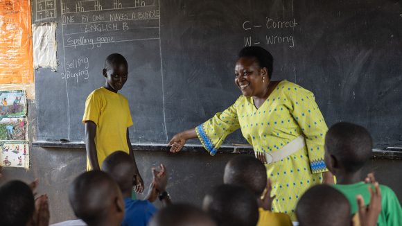 A female teacher stands in front of a chalk board in a classroom. She is smiling and laughing with students.