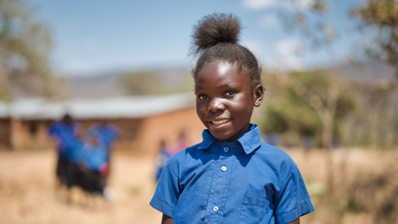 A girl wearing a blue shirt smiles while standing in a school playground.