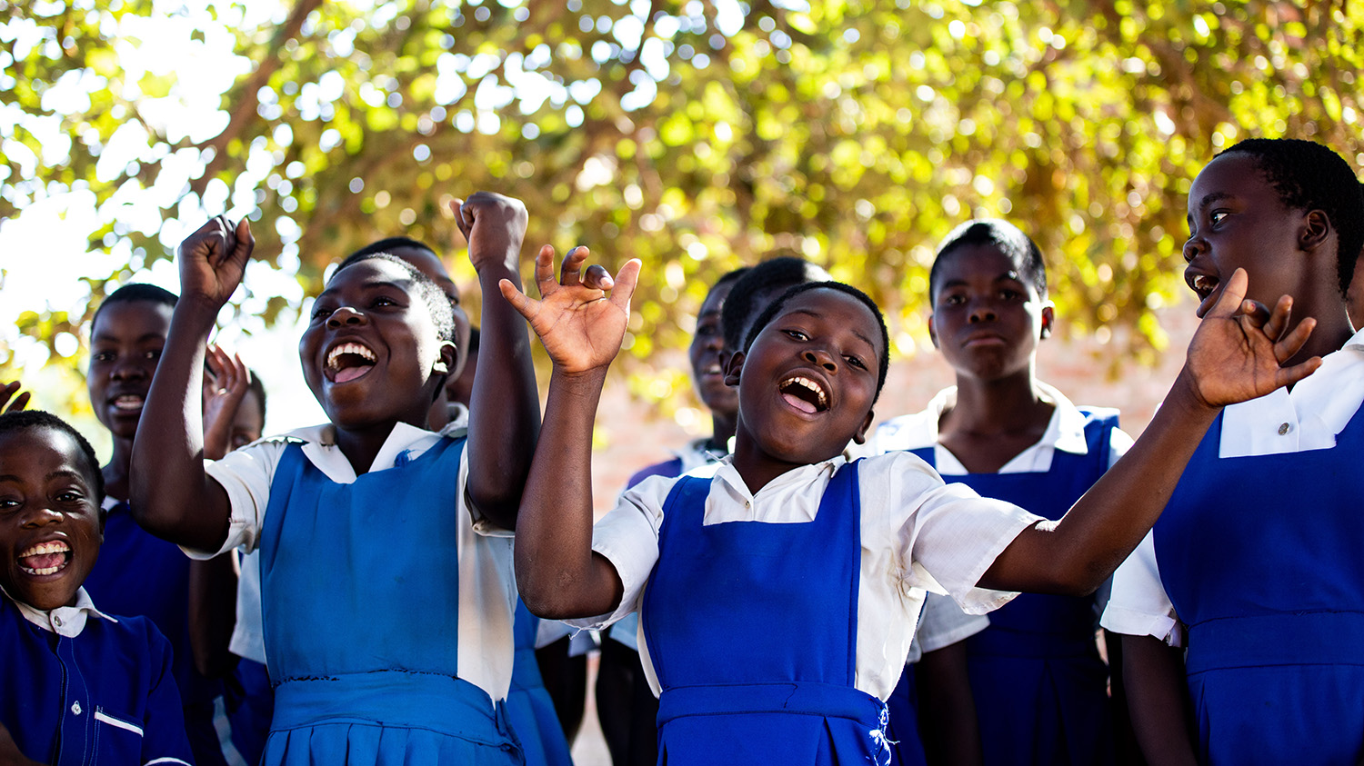 A group of teenage girls wearing school uniform cheer and wave their arms in the air.