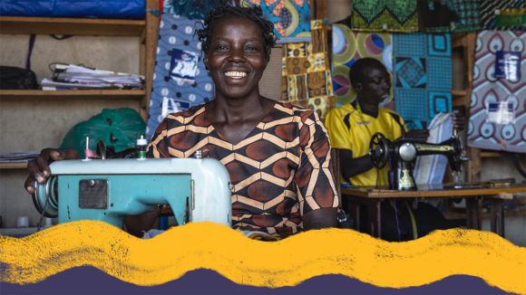 A woman smiles broadly while sitting at a sewing machine. Behind her, a man is inspecting a piece of fabric.