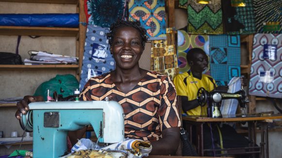 A woman smiles broadly while sitting at a sewing machine. Behind her, a man is inspecting a piece of fabric.