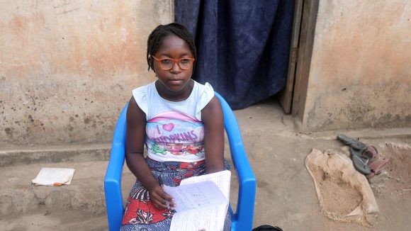 A girl wearing a pair of glasses sits in a chair outside her home. She is writing in an exercise book.