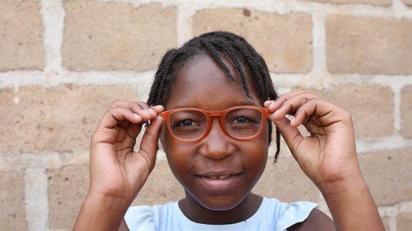 A girl wearing a red pair of glasses stands in front of a brick wall.