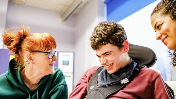 Two women and a teenage boy laugh and chat. The teenager is using an electric wheelchair and wearing a safety harness.