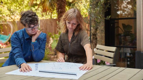 Two women review design plans at a table in a leafy garden.