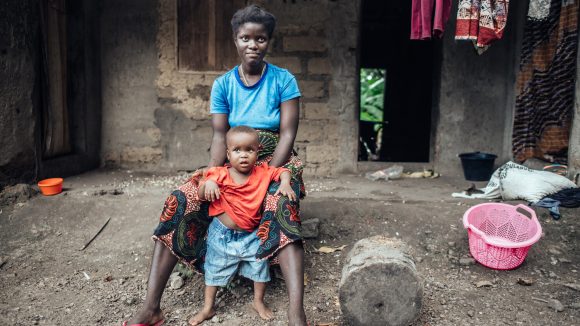 Susan sits outside her home holding her young son, Yusif. The floor is rough dirt, and there's washing hanging in the background.