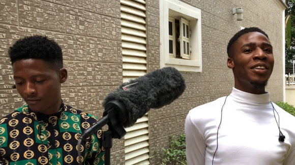 A man with earphones around his neck smiles while another man holds a boom mic up to him.