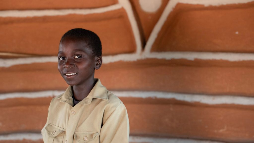 11 year old Twinde outside his home in Zimbabwe, smiling at the camera.