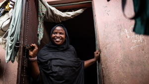 A woman wearing a head scarf smiles broadly while standing in a doorway.