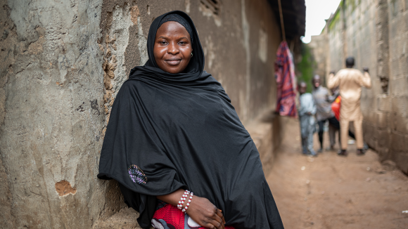 A woman wearing a black headscarf and tunic smiles while leaning against a wall.