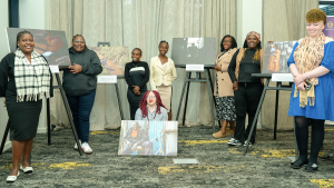 Eight women proudly smile beside large canvases on easels.