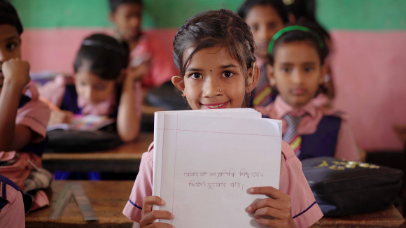 A young girl wearing a hearing aid smiles behind a stack of papers at a classroom desk.