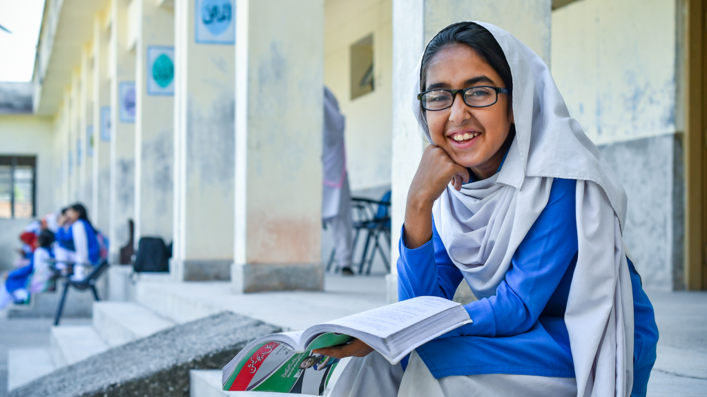 Minahil sits down outside at school with a book. She rests her head on her hand and looks at the camera.