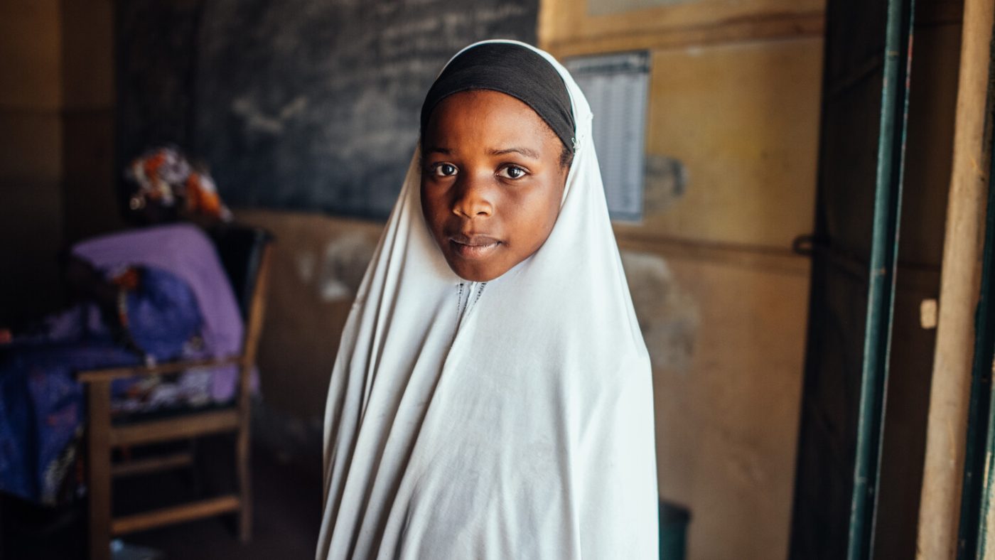 Rukayya wears a white head covering while standing in a classroom.