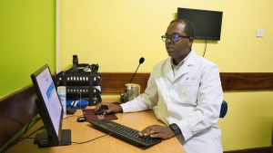 A man wearing a white medical coat sits in front of a computer at a desk.